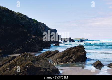 Une vue panoramique sur la côte avec des falaises rocheuses et une plage de sable. Les vagues s'écrasent doucement contre les rochers sombres, avec un ciel bleu clair et des collines lointaines dedans Banque D'Images