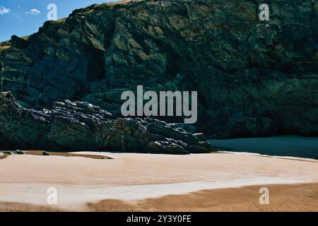 Une scène de plage sereine avec sable doré lisse et rochers sombres accidentés. La toile de fond se compose d'une falaise rocheuse abrupte avec un mélange de textures et de couleurs Banque D'Images