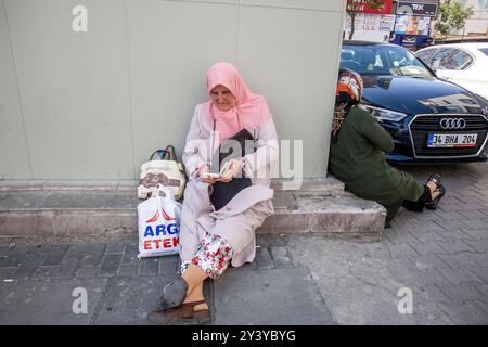 Ankara, Turquie - 13 juin 2022 : une femme vêtue d'un hijab rose est assise sur le trottoir avec ses effets personnels, tandis qu'une autre repose contre un coin de bâtiment. Banque D'Images