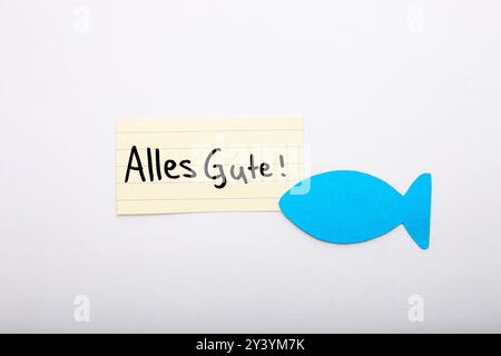 Alles Gute ! Mot de souhait allemand pour la communion sur un papier beige, écrit à la main. Isolé sur blanc. Espace libre pour le texte. Poisson en papier bleu dans le coin. Banque D'Images