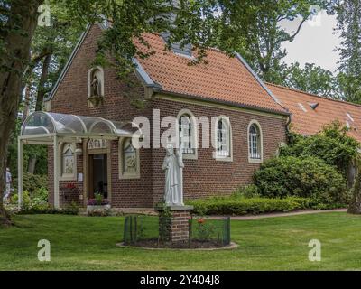 Une petite église en briques avec des tuiles rouges, entourée d'un jardin verdoyant avec une statue au premier plan, Reken, Muensterland, Allemagne, Europe Banque D'Images