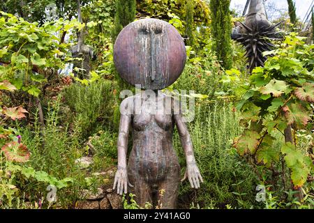 Une sculpture unique d'une figure humanoïde avec une grande tête ronde et un design minimaliste, entourée de verdure luxuriante et de plantes dans un cadre de jardin. Banque D'Images