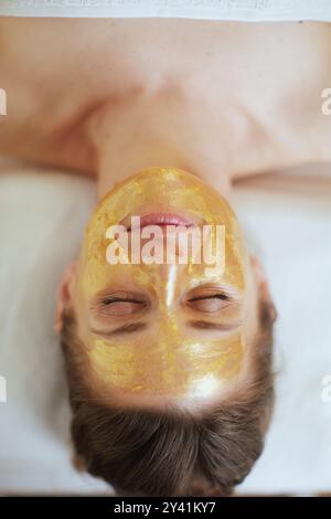 Temps de soins. Vue supérieure de la femme moderne d'âge moyen détendue dans le salon de spa avec masque cosmétique doré sur le visage couché sur la table de massage. Banque D'Images