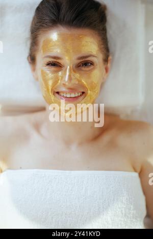 Temps de soins. Vue supérieure de femme moderne souriante dans le salon de spa avec masque cosmétique doré sur le visage couché sur la table de massage. Banque D'Images