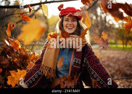 Bonjour l'automne. jeune femme souriante en chapeau rouge avec écharpe jetant des feuilles d'automne dans le parc de la ville. Banque D'Images