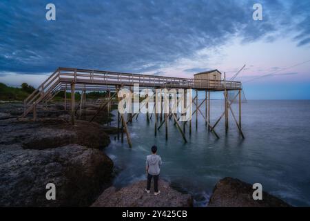 Paysages de coucher de soleil sur la côte de Saint-palais-sur-mer, avec un homme debout sur la falaise regardant les carrelets traditionnels le long de la mer à Cha Banque D'Images