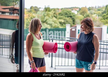 Deux femmes plus âgées, vêtues de vêtements de sport, se tiennent debout à l'entrée du gymnase, discutant devant leur classe, avec des tapis de yoga cachés sous leurs bras Banque D'Images