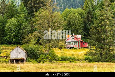 Vieille maison rouge dans un champ entouré de grands arbres, avec un hangar de bateau plus petit à proximité. Côte nord de la Colombie-Britannique sur l'île Porcher. Banque D'Images