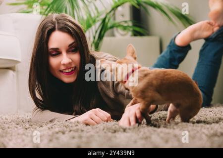 Deux femmes sont allongées sur un tapis, jouant joyeusement avec leur petit chien brun. Une femme caresse le chien pendant que l'autre regarde Banque D'Images