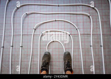 Vue de dessus sur les chaussures de travailleurs comme debout sur de minces tubes blancs courbés dans un collecteur de système sous plancher complexe monté pour le plancher de chauffage central dans un resi Banque D'Images