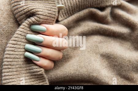 Une main féminine avec de longs ongles verts élégants tient doucement un pull beige doux, mettant en valeur une esthétique confortable et à la mode parfaite pour l'automne. Banque D'Images