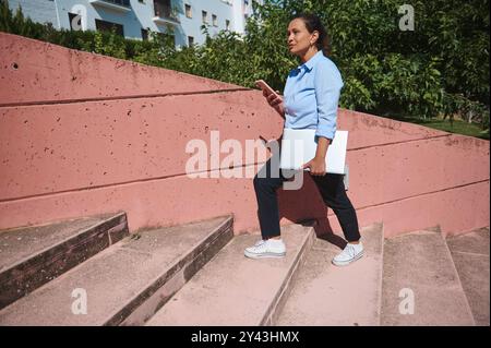 Une femme professionnelle monte les escaliers, tenant un ordinateur portable et utilisant son smartphone, mettant en valeur le multitâche et l'entreprise moderne. À l'extérieur avec de la verdure, s Banque D'Images