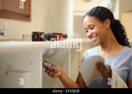 Femme noire heureuse installant le manuel de mobilier dans la cuisine Banque D'Images