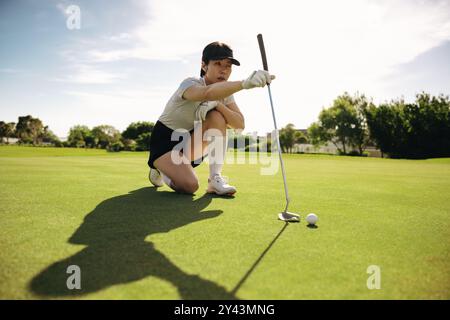 Golfeuse concentrée s'agenouillant et alignant un putt sur une journée ensoleillée et lumineuse à l'extérieur sur un parcours de golf luxuriant. Banque D'Images