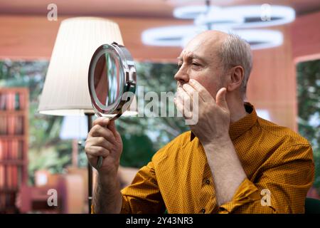 Homme senior regardant son visage vieillissant dans le miroir et essayant de préserver sa peau, en utilisant l'hydratation et le sérum de protection UV. Personne âgée appliquant des produits de toilettage sur une peau vieillissante. Banque D'Images