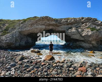 Personne debout dans l'océan regardant à travers la voûte rocheuse naturelle sur une plage. Banque D'Images