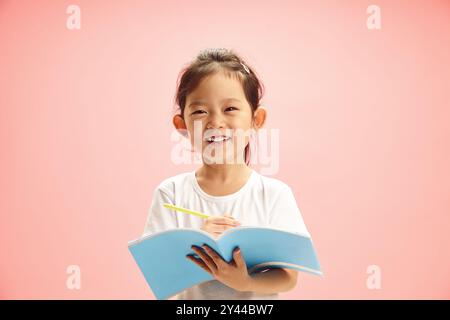 Jeune fille chinoise de première année joyeuse souriante tout en tenant un cahier d'exercices et un stylo debout sur rose isolé Banque D'Images