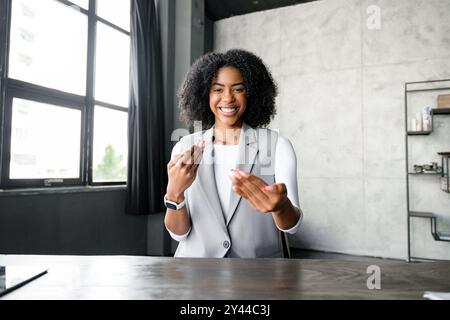Une femme afro-américaine participe à une conversation virtuelle, ses gestes animés s’ajoutant à l’atmosphère dynamique du bureau moderne, parfait pour illustrer le côté humain du travail à distance Banque D'Images