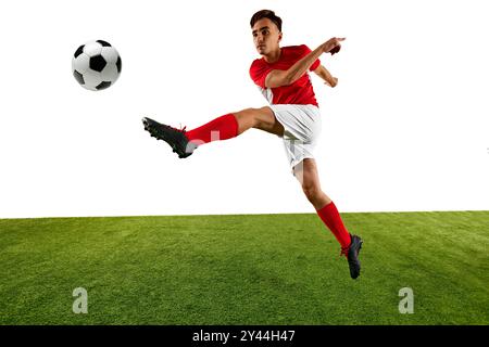 Jeune homme athlétique, joueur de football, vêtu d'un uniforme rouge et blanc, effectue un coup de pied élevé, en se concentrant sur le contrôle du ballon avec précision sur le terrain vert. Banque D'Images