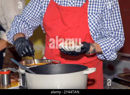 Chef vêtu d'un tablier rouge cuisine, tenant des tranches de saucisse, des ingrédients assidus, préparant un plat délicieux dans une cuisine professionnelle, arts culinaires c Banque D'Images