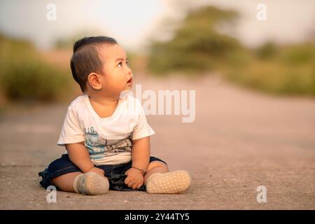 mignon bébé asiatique assis sur la route. bébé garçon tourné sur le côté portant des vêtements blancs. ambiance photo dans l'après-midi. Banque D'Images