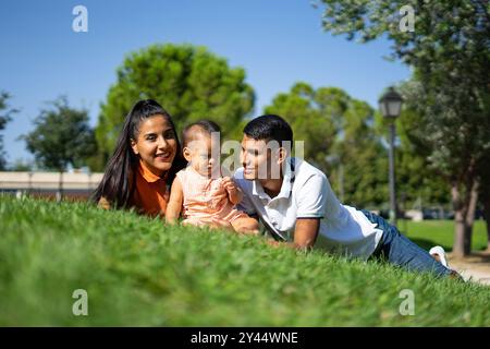 Jeune famille latine de père, mère et petite fille allongée sur l'herbe d'un parc par une journée ensoleillée Banque D'Images