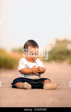 mignon bébé asiatique assis sur la route. bébé garçon tenant de l'herbe sauvage tout en souriant portant des vêtements blancs. ambiance photo dans l'après-midi. Banque D'Images