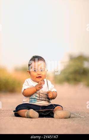 mignon bébé asiatique assis sur la route. bébé garçon tenant de l'herbe sauvage tout en souriant doucement portant une chemise blanche vers la caméra. ambiance photo en t Banque D'Images