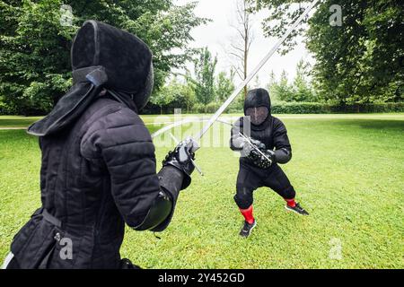 Deux escrimeurs vêtus d'uniformes de protection noirs, des casques avec des masques faciaux sont des clôtures dans le parc de la ville en utilisant de longues épées historiques en métal médiéval, practi Banque D'Images