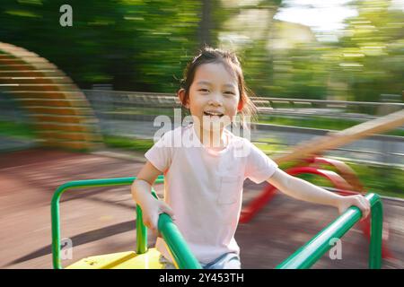 Jeune fille asiatique joyeuse de cinq ans souriante tout en chevauchant Playground Merry-Go-Round Banque D'Images