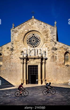 Deux cyclistes pédalent devant la façade de la cathédrale romane d'Otrante. La basilique catholique date du 11ème siècle et se trouve à Otrante, dans les Pouilles, Banque D'Images