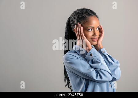 Jeune femme afro-américaine souriante avec de longs cheveux tressés regardant loin tout en touchant les joues contre le mur gris Banque D'Images