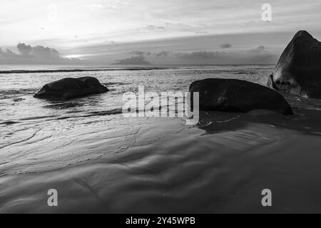 Photographie de paysage noir et blanc avec sable humide et silhouettes de rochers côtiers sur le coucher du soleil. Plage de beau Vallon, île de Mahé, Seychelles Banque D'Images