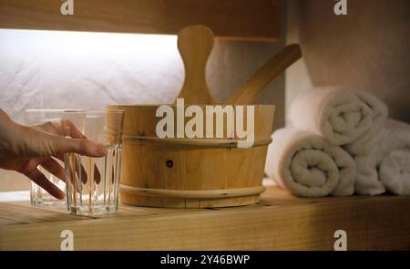 Gros plan d'une main d'une femme tenant un verre d'eau debout sur un banc en bois dans un sauna, seau de sauna avec louche et une pile de trois serviettes roulées Banque D'Images