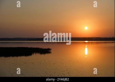 Cette photo sereine capture la beauté d'un coucher de soleil sur un lac calme, avec le reflet du soleil scintillant sur l'eau. L'horizon est bordé de t Banque D'Images