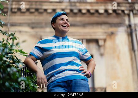 Individu joyeux de genre non conforme avec les cheveux bleus courts et chemise rayée riant à l'extérieur. Expression authentique du bonheur et de la confiance en soi Banque D'Images