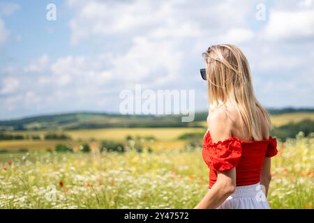 Jeune femme blonde en lunettes de soleil dans une robe blanche rouge dans un champ de fleurs sauvages Banque D'Images