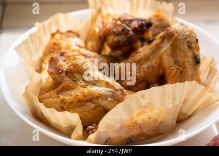 Les ailes de poulet fraîchement frites sont placées sur du papier dans une assiette blanche avec la lumière brillante à travers la fenêtre tombant sur le poulet. Repas fait maison cuisiné dans le gril de la friteuse à air. Mise au point sélective. Banque D'Images