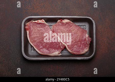 Steaks de porc crus couchés dans un plateau en plastique noir sur fond sombre vue de dessus Banque D'Images