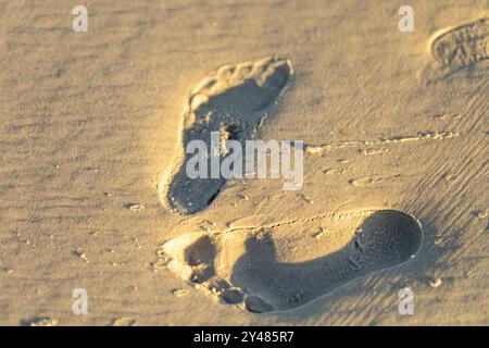 Deux empreintes de pas sont gravées dans le sable chaud, luisant doucement sous la lumière du soleil couchant, reflétant une atmosphère paisible à la plage. Banque D'Images