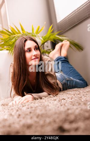 La jeune femme est confortablement allongée sur le ventre sur un tapis moelleux dans son salon confortable, se prélasser dans la lumière naturelle. Elle aime une journée paisible à la maison, Fee Banque D'Images