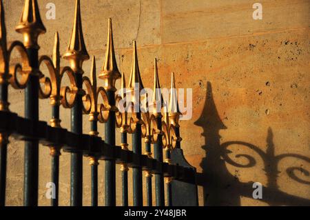 Élégante clôture en fer avec des pointes dorées jetant des ombres sur le mur de pierre ensoleillé Banque D'Images