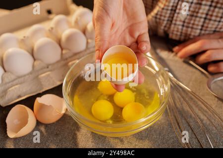 Une personne cassant un oeuf dans un bol en verre, avec plusieurs oeufs dans un carton d'oeufs en arrière-plan. La scène se déroule dans une cuisine, mettant en valeur les processus Banque D'Images