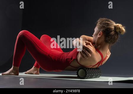 Femme effectuant la libération myofascial des muscles hyperélastiques du dos avec un rouleau de massage sur un fond sombre. Concept de prévention de la fatigue dorsale et se Banque D'Images