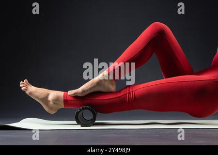 massage de physiothérapie, femme avec rouleau de yoga sur le sol pour la tension des jambes sur fond sombre, auto-massage avec rouleau Banque D'Images