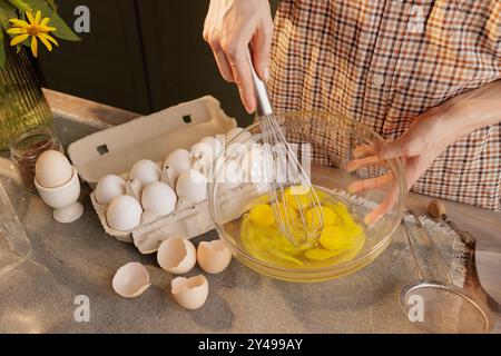 Gros plan de mains féminines battant des œufs avec un fouet à main dans un bol transparent sur le fond d'une table de cuisine. Oeufs dans une boîte et un outil pour Banque D'Images