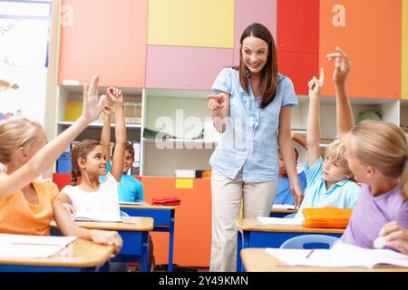 Femme, question et enfants avec les mains levées en classe pour l'éducation, la discussion et le choix de la réponse. Heureux, professeur et étudiants avec geste à Banque D'Images
