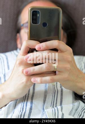 Un homme regarde son téléphone tout en étant allongé sur un canapé. Il porte une bague de mariage et le téléphone couvre son visage Banque D'Images