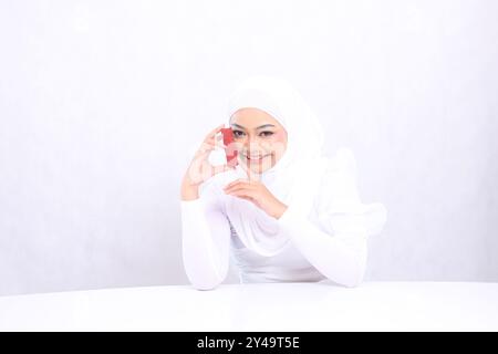 gros plan sur le maquillage des lèvres. Belle jeune fille de voile joyeuse regardant la caméra. Main tenant et touchant le récipient de rouge à lèvres isolé sur fond blanc Banque D'Images