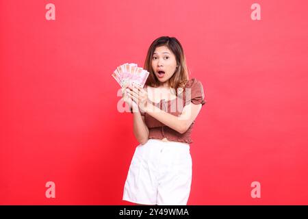 l'expression de la femme indonésienne choquée, les deux mains tenant de la monnaie rupiah, portant une blouse brune, fond rouge. pour la transaction, la technologie et adve Banque D'Images